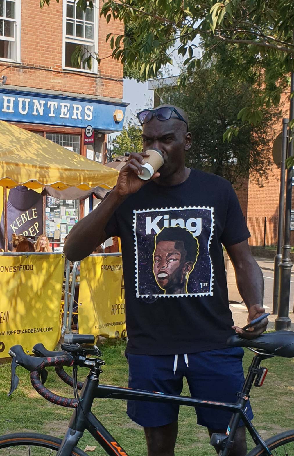 black man drinking from a cup while standing next to a bicycle, wearing a t-King tshirt with a graphic design.