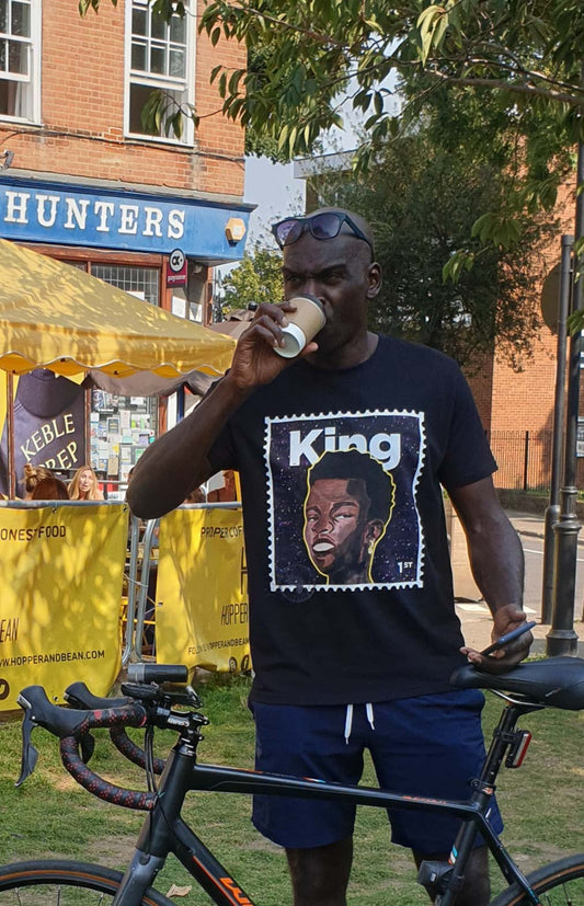 black man drinking from a cup while standing next to a bicycle, wearing a t-King tshirt with a graphic design.