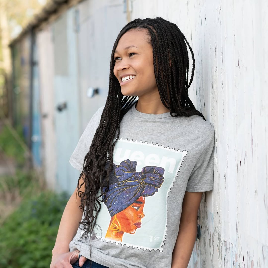 Woman wearing a gray t-shirt with a colourful graphic design of a black queen, leaning against a white wall.