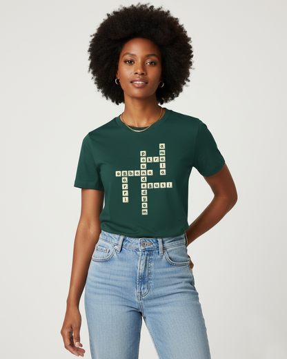 Black woman in her early 30s, with a stylish afro, stands confidently against an off-white studio background. She is wearing a dark green t-shirt with a unique crossword puzzle design on the front, featuring Nigerian food names