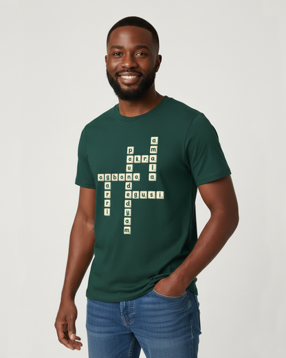 Black man in his early 30s, with a stylish afro, stands confidently against an off-white studio background. He is wearing a dark green t-shirt with a unique crossword puzzle design on the front, featuring Nigerian food names